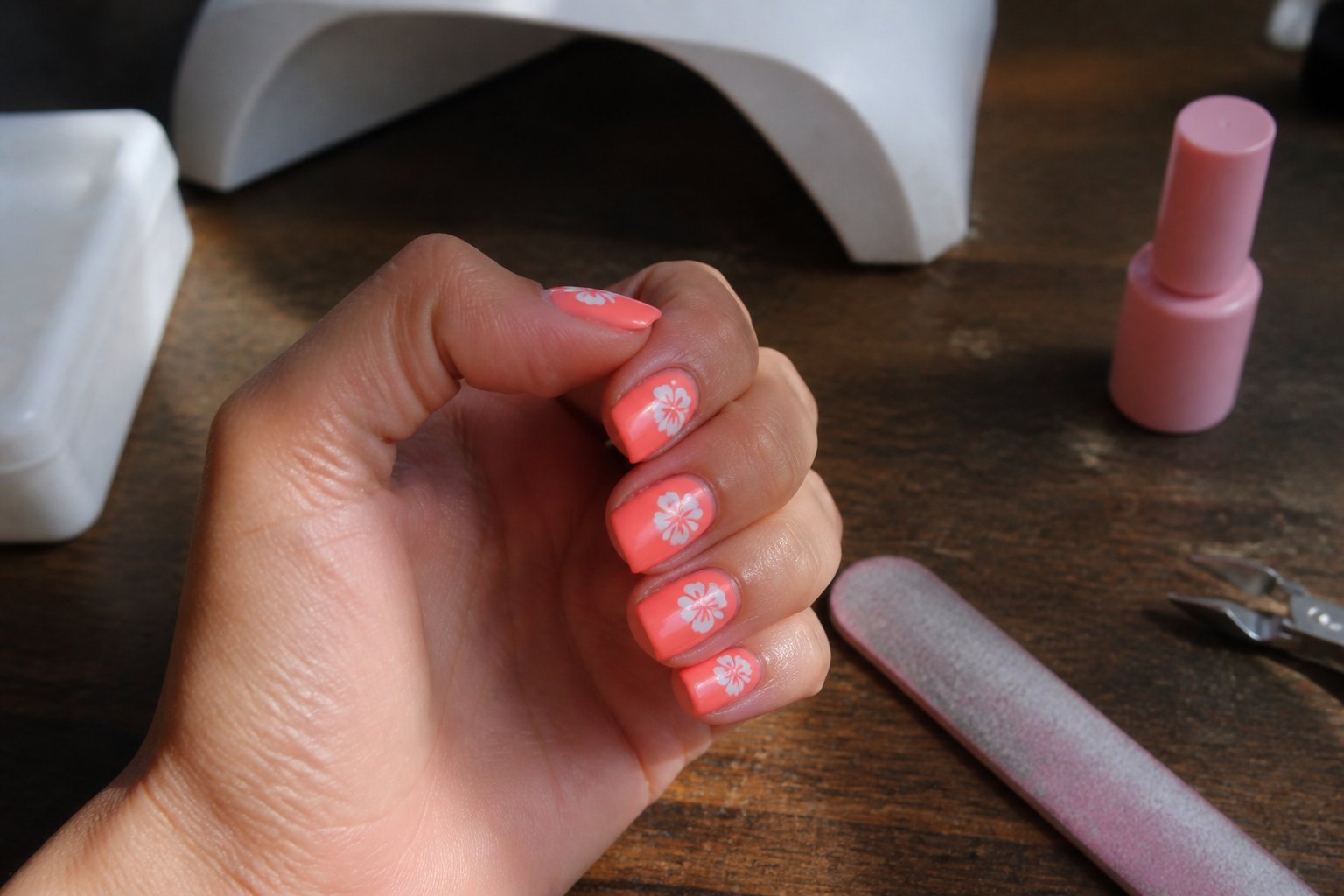 Close-up of hands with short neon coral coffin-shaped nails featuring tiny white hibiscus accents, resting on a wooden surface with manicure tools nearby.