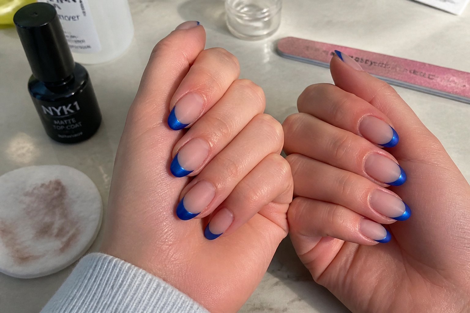 Close-up of hands with cobalt blue French tip nails in matte finish, surrounded by manicure tools on a softly lit surface.