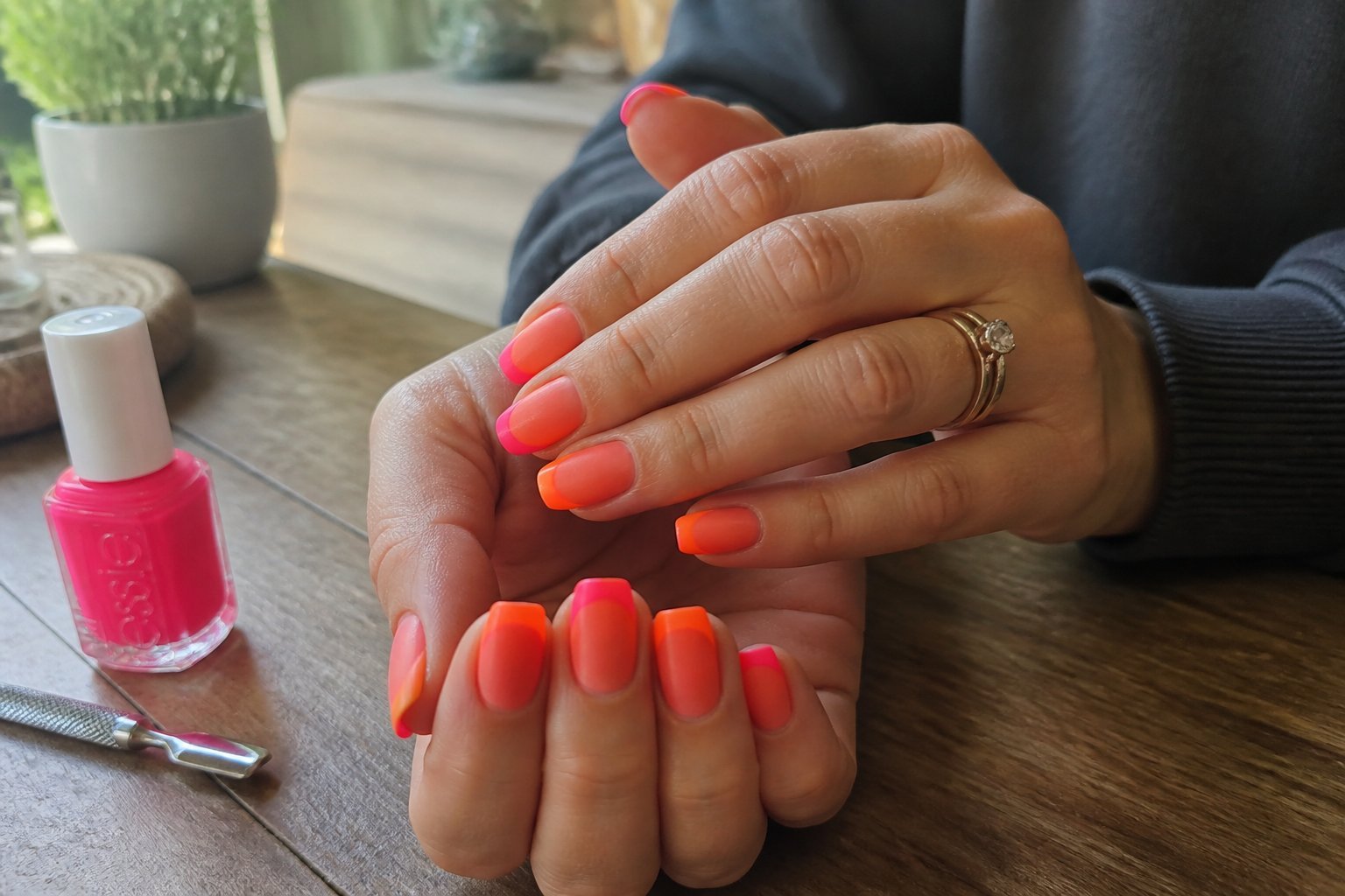 Close-up of hands with neon French tip nails featuring a gradient orange-to-pink base resting on a wooden surface with manicure tools nearby.