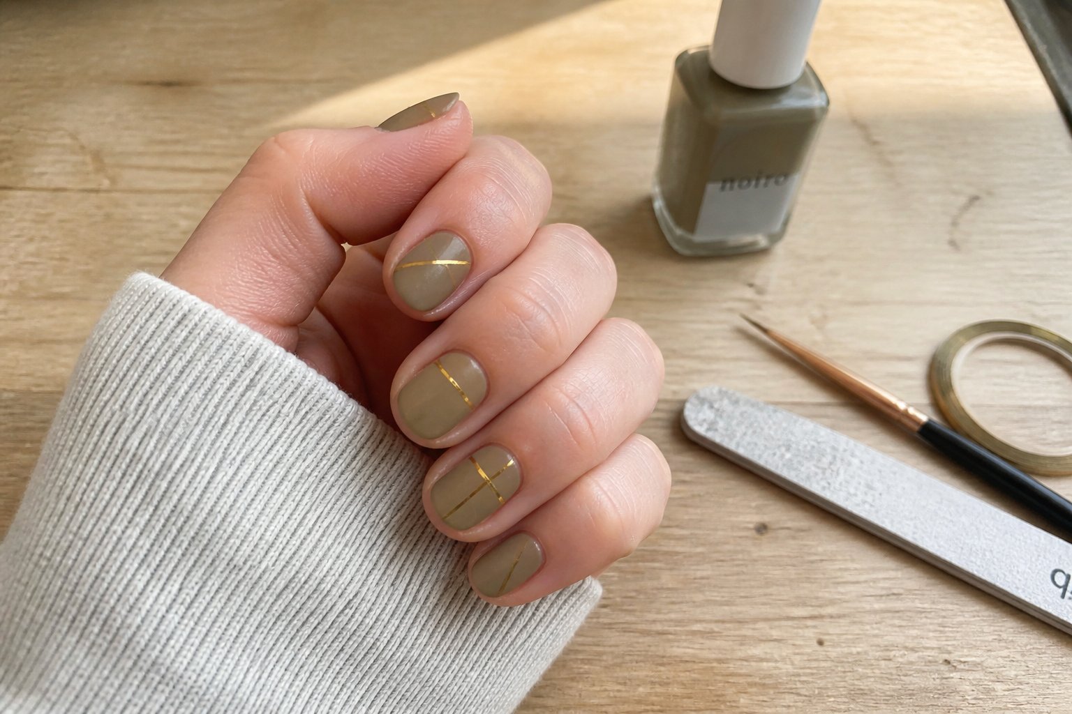 Close-up of hands with soft khaki nails and thin gold stripes, surrounded by manicure tools on a wooden surface.