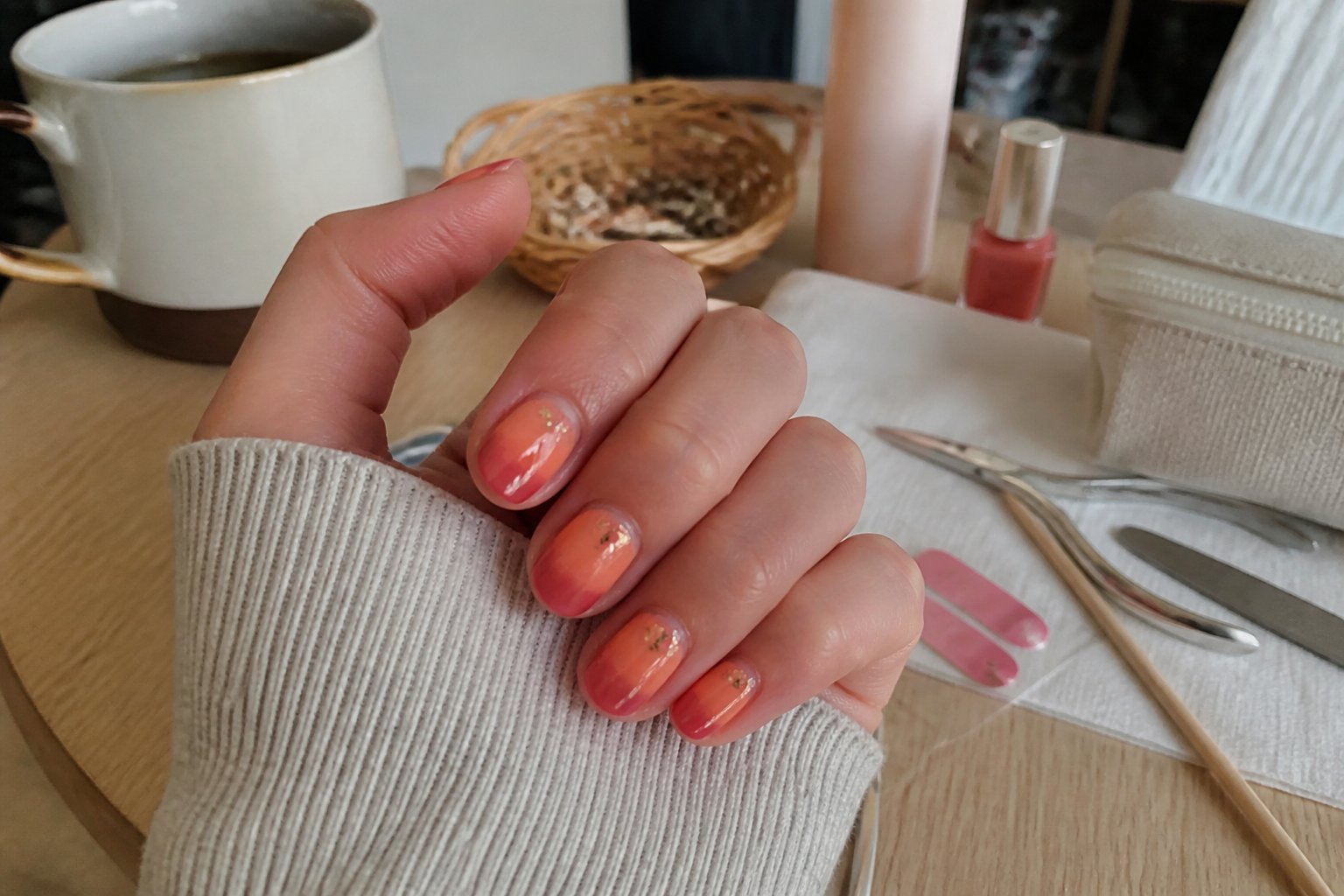 Hands with sunset ombre nails in coral, peach, and gold flakes surrounded by manicure tools on a softly lit surface.