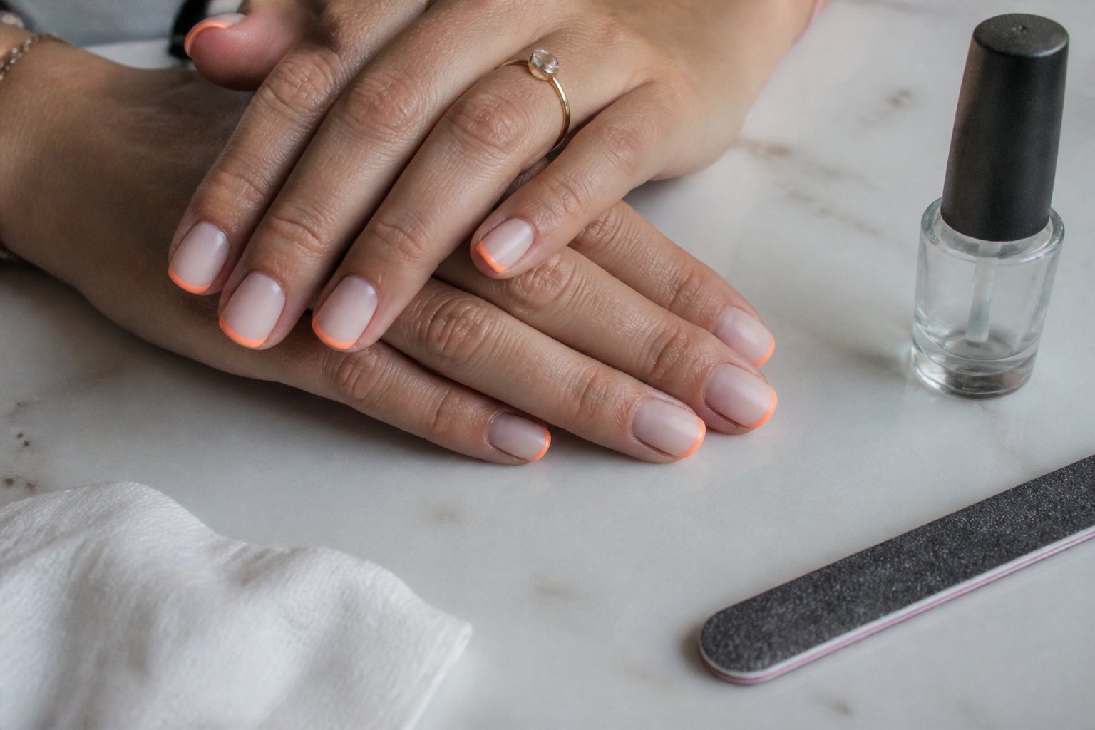 Close-up of hands with white French manicure and thin neon orange tips, resting among nail care tools on a light surface.