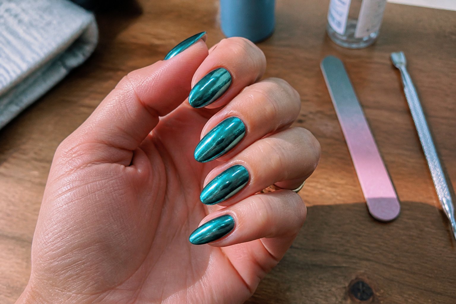 Close-up of hands with teal chrome almond-shaped nails resting near manicure tools on a wooden surface.