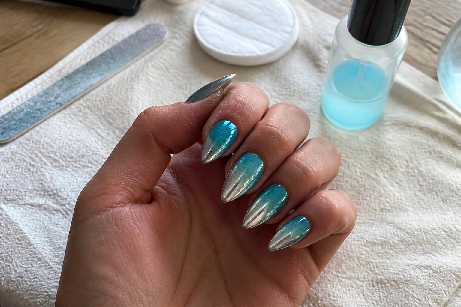 Close-up of a hand with teal-to-silver ombre chrome stiletto nails resting on a wooden surface, surrounded by manicure tools.