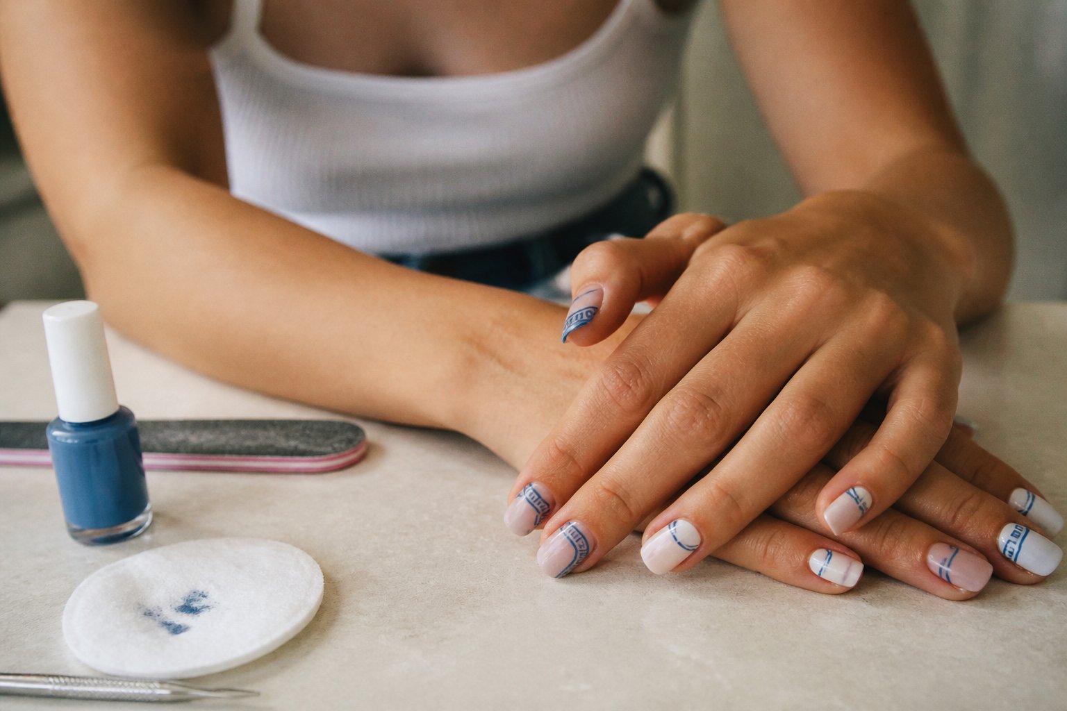 Close-up of hands with blue and white Mediterranean-inspired nail art surrounded by manicure tools on a matte surface in soft natural daylight.