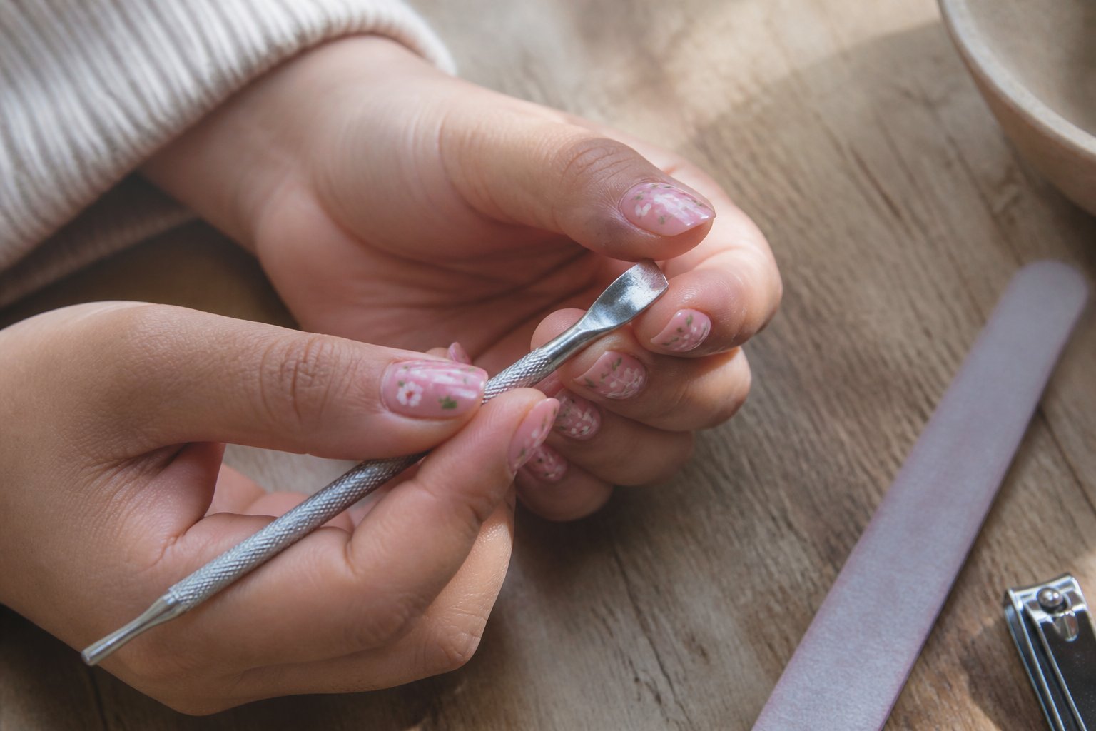 Close-up of hands with soft pink nails featuring tiny floral patterns, holding nail tools on a textured surface.