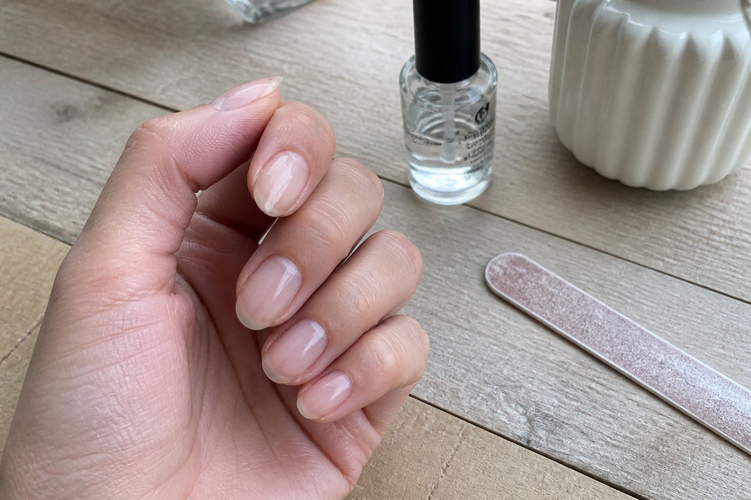 Close-up of a hand with sheer beige ombré nails resting on a wooden surface next to manicure tools.