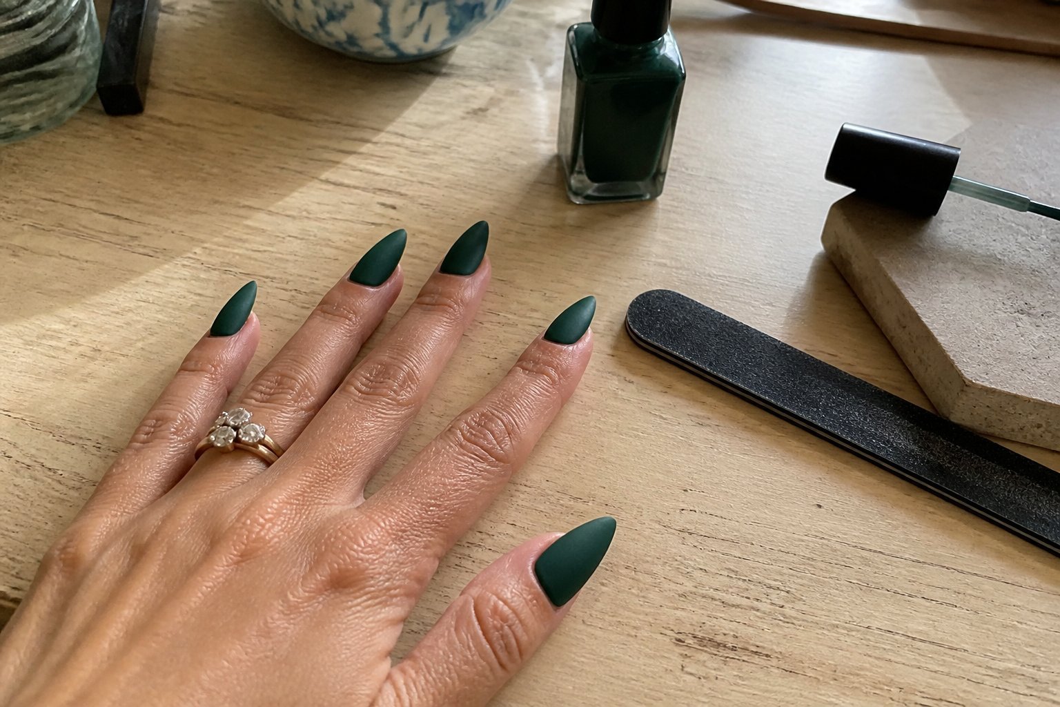 A hand with deep emerald jade matte stiletto nails resting on a wooden table with manicure tools nearby.