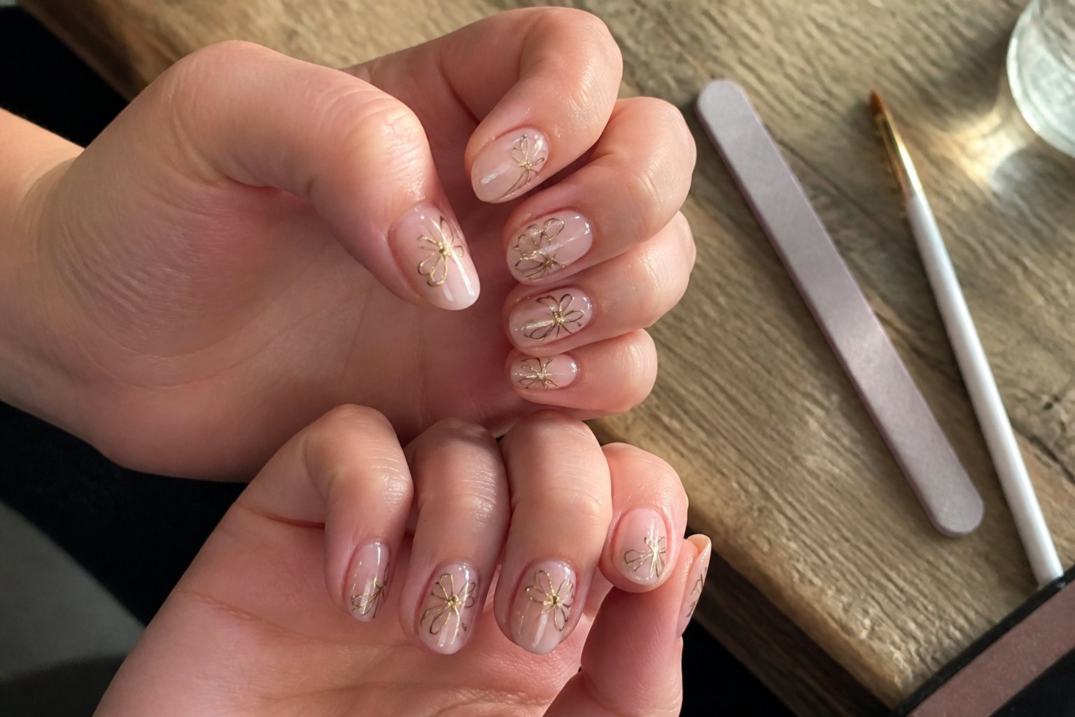 Close-up of hands with nude nails featuring gold-outlined dragonfly accents, resting near manicure tools on a wooden surface.