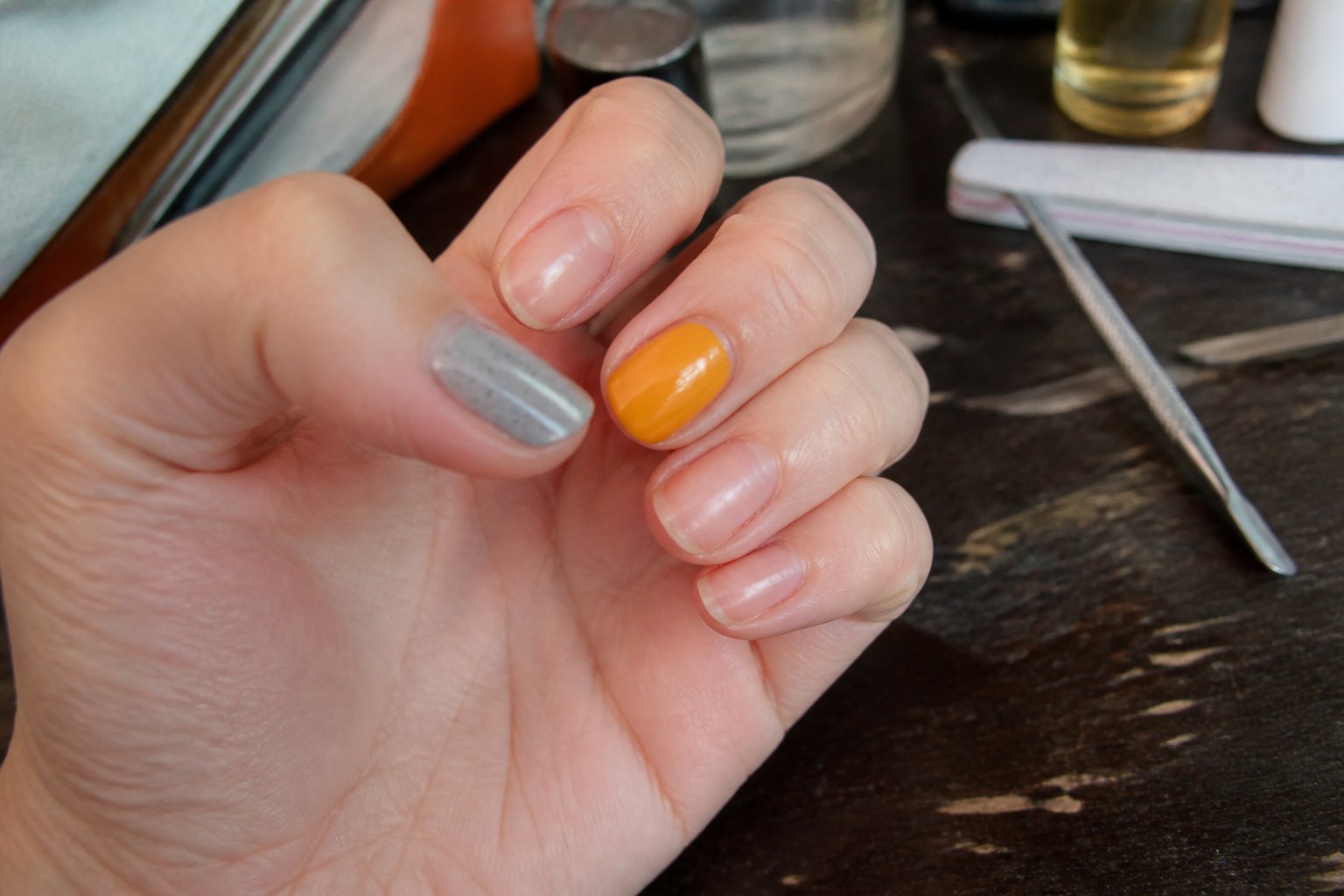Close-up of a hand with mismatched nails: a speckled gray thumb and a glossy marigold middle finger, resting on a clean surface with some manicure tools nearby.