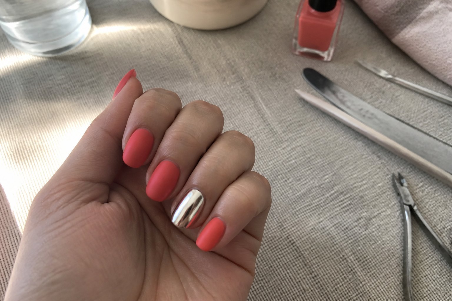 A hand with one chrome silver nail and the other nails painted matte coral resting on a textured surface with manicure tools nearby.