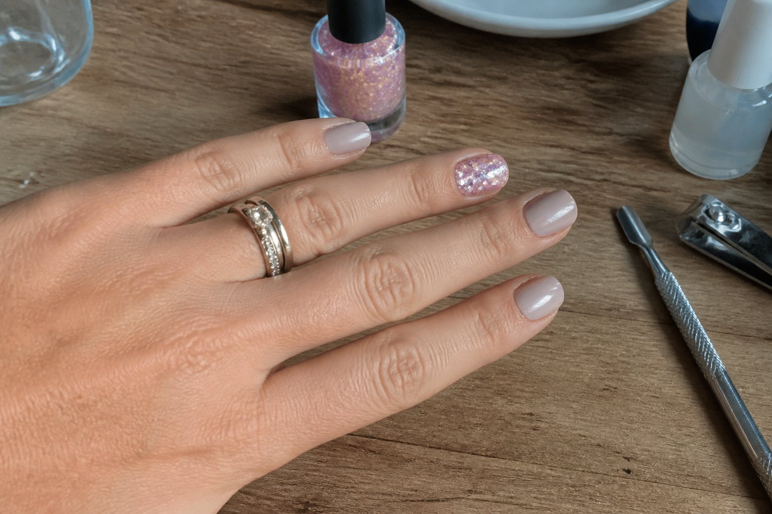 Close-up of a hand with holographic pink nail polish on the ring finger and satin beige on the other nails, resting on a wooden table with manicure tools nearby.