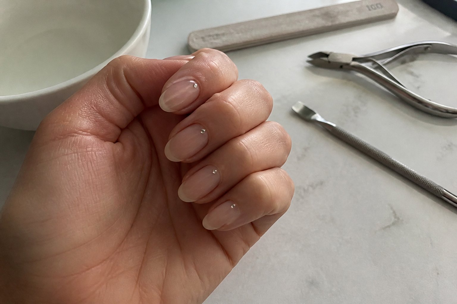 Close-up of a hand with translucent beige micro French nails and tiny rhinestones near the cuticles, resting on a table with manicure tools nearby.