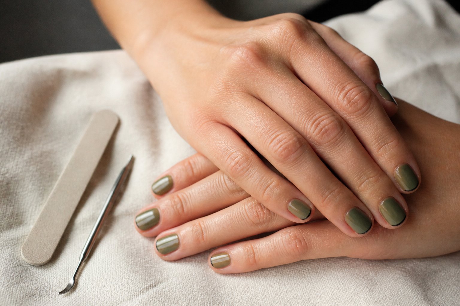 Close-up of hands with olive green micro French manicure with thin black outlines resting on a wooden surface with manicure tools nearby.