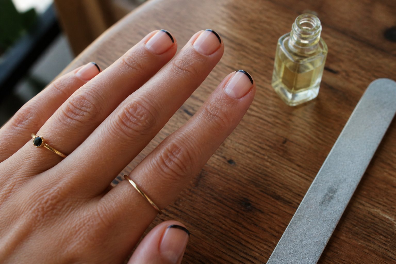 Close-up of short square nails with a delicate black micro French manicure on a hand resting on a wooden surface with nail care tools nearby.