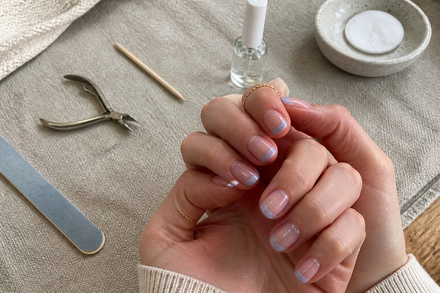 Hands with pearlescent baby blue micro french nails with glitter fade, surrounded by manicure tools on a textured surface.