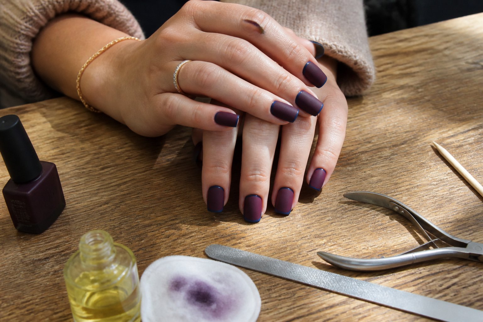 Close-up of hands with plum noir square nails edged in midnight blue resting on a wooden table with manicure tools nearby.