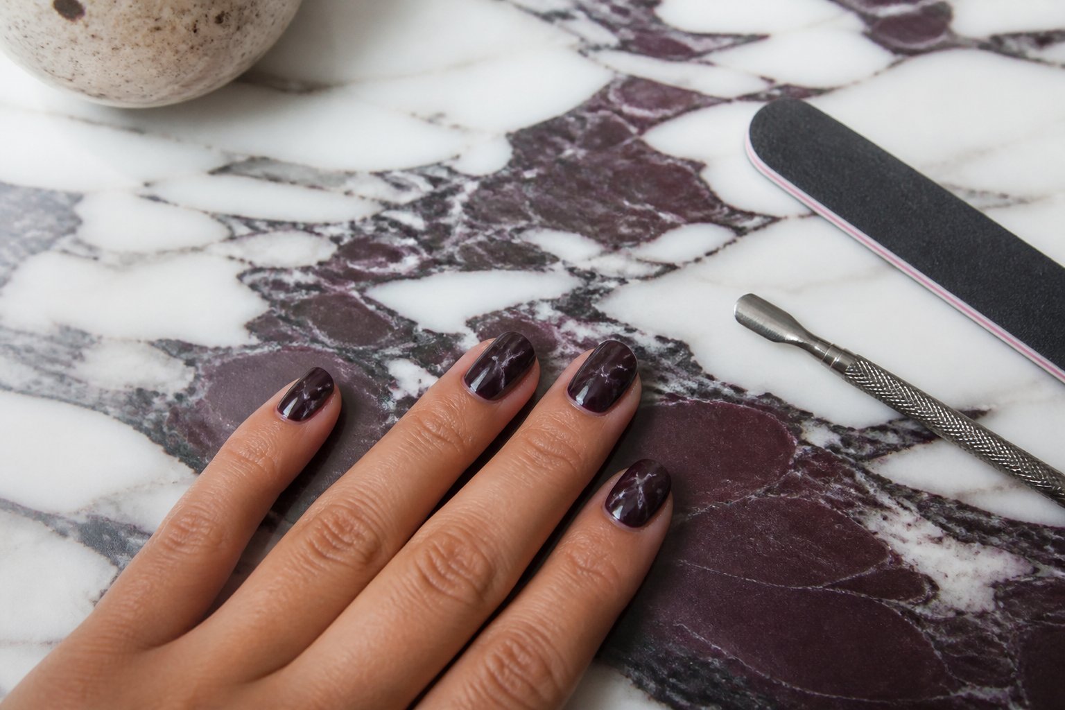 A hand with plum noir nails featuring pale taupe vein accents resting on a marble surface with manicure tools nearby.