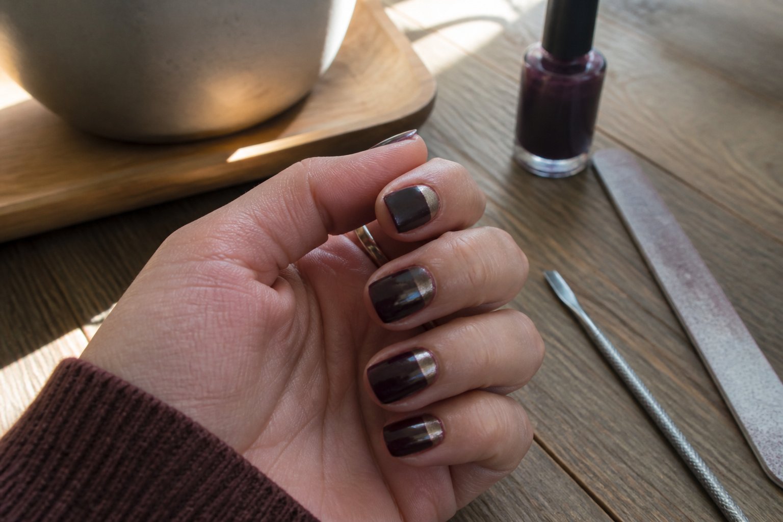 Close-up of a hand with plum noir stiletto nails featuring a pearl chrome half-moon design resting on a wooden surface with manicure tools nearby.
