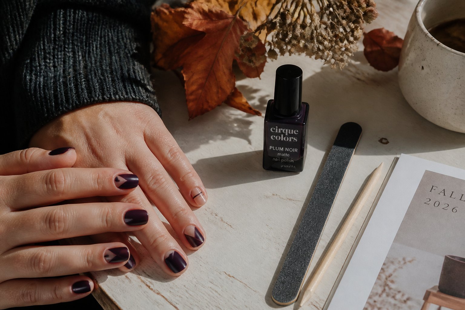 Hands with Plum Noir nails featuring geometric designs on a wooden table with manicure tools and soft daylight.