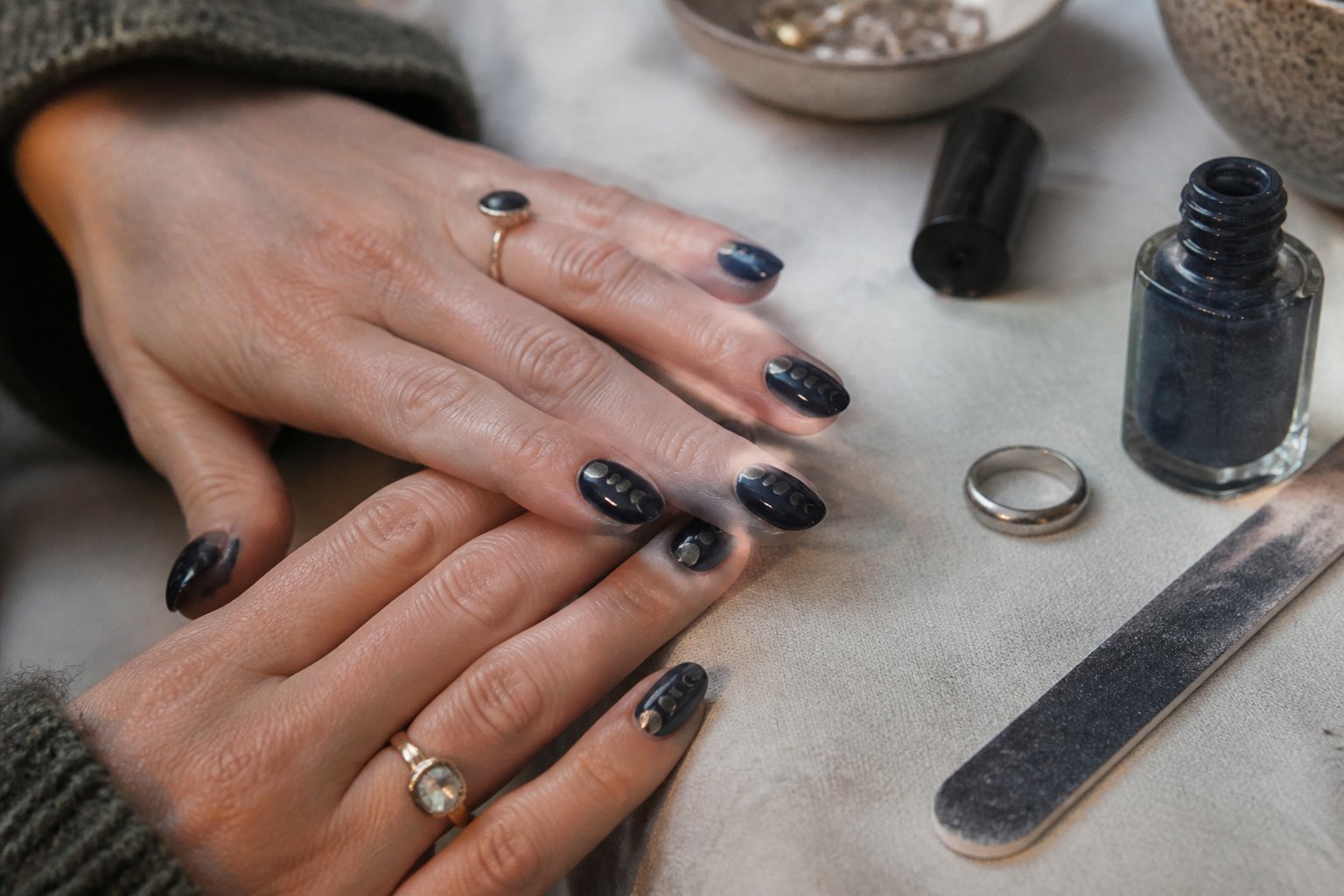 Close-up of hands with midnight navy jelly nails decorated with silver moon phase decals, surrounded by nail care tools on a softly lit surface.