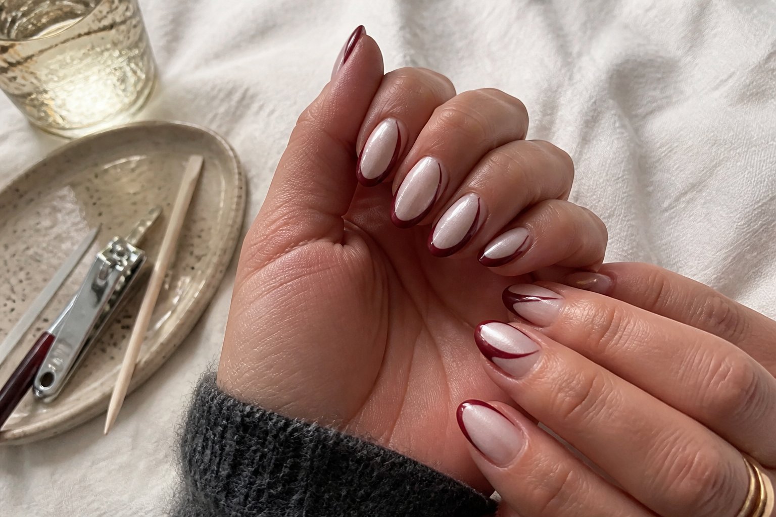 Close-up of hands with opalescent pearl nails and dark red French tip reverse smirk design resting on a soft fabric surface with manicure tools nearby.