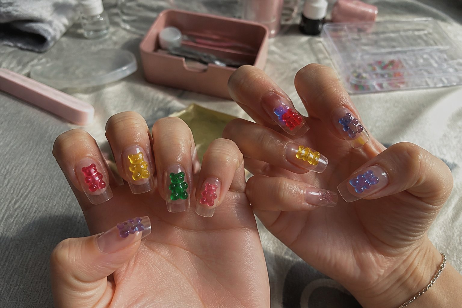Close-up of hands with clear nails decorated with tiny gummy bear designs, surrounded by manicure tools on a softly lit surface.