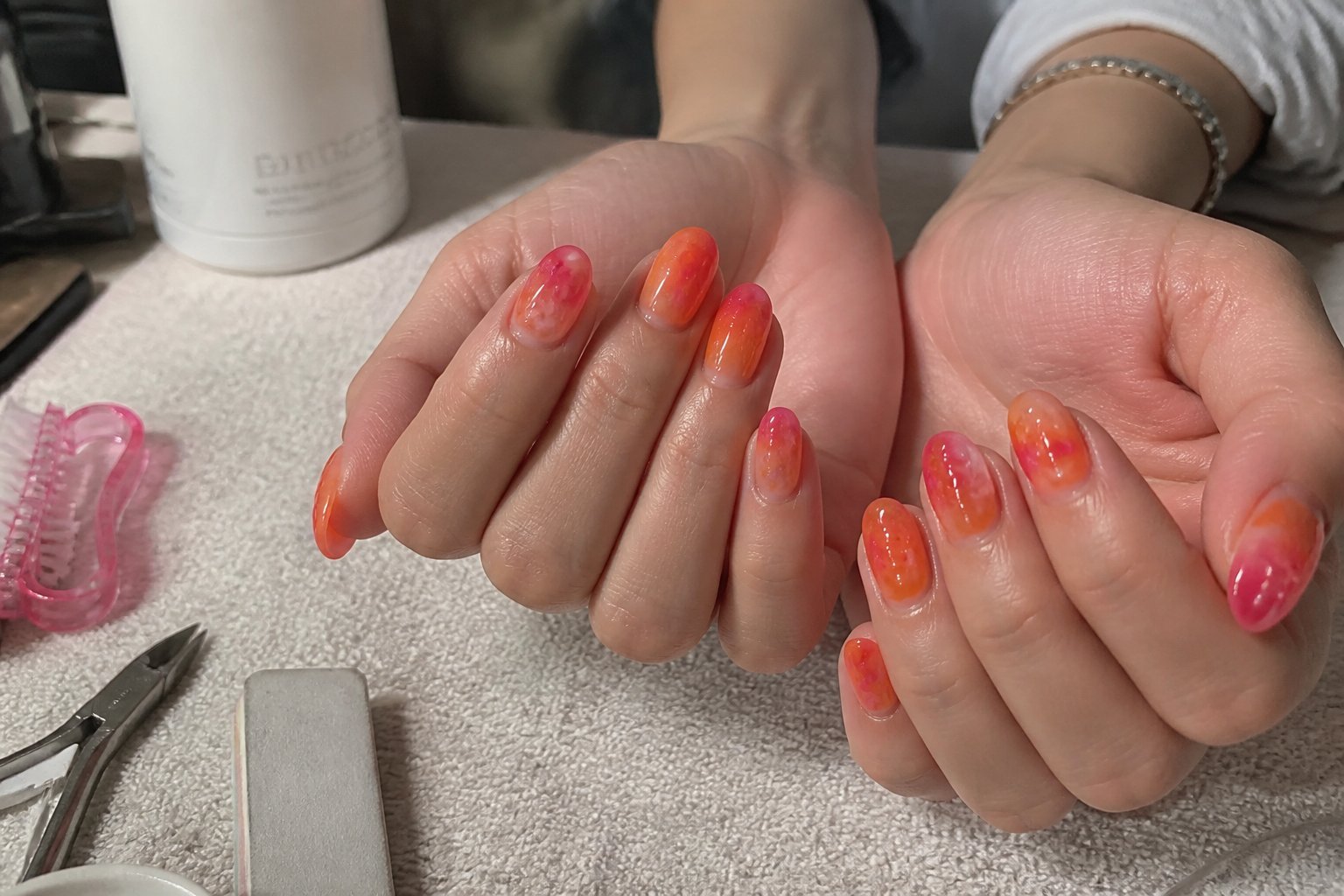 Close-up of hands with neon orange to hot pink gradient jelly nails resting near nail tools on a textured surface in natural daylight.