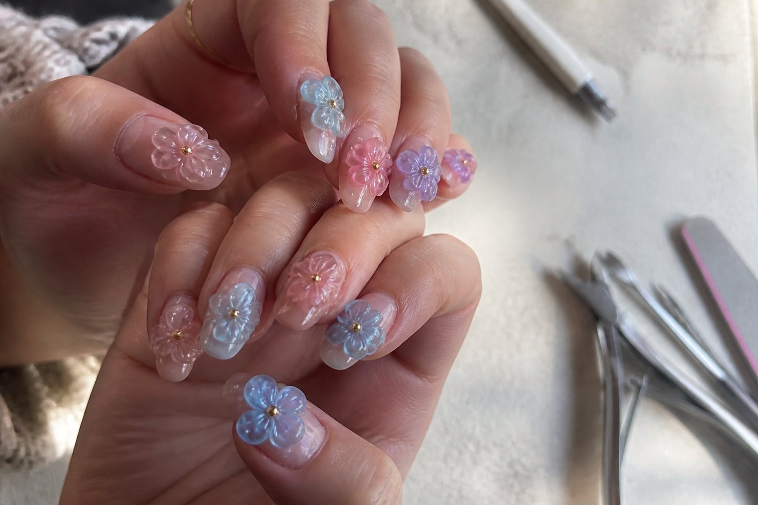 Close-up of hands with translucent 3D flower jelly nails and raised petal gel, surrounded by manicure tools on a simple surface.