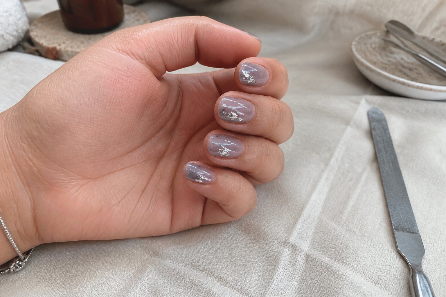 A close-up of a hand with short smoky grey jelly nails decorated with silver foil flakes resting on a wooden surface with nail care tools nearby.