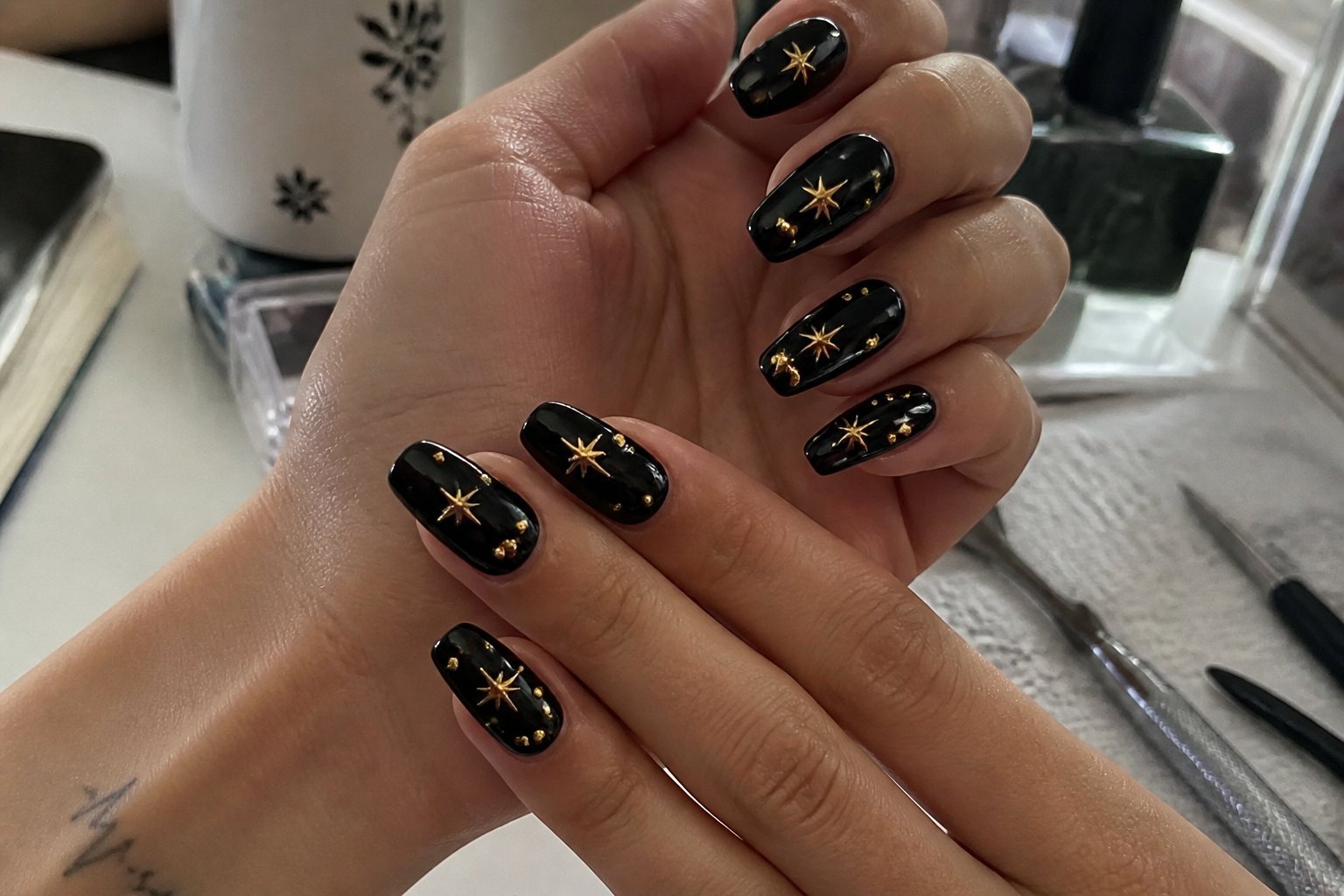 Close-up of hands with black jelly nails decorated with tiny gold star charms, surrounded by manicure tools on a casual surface.